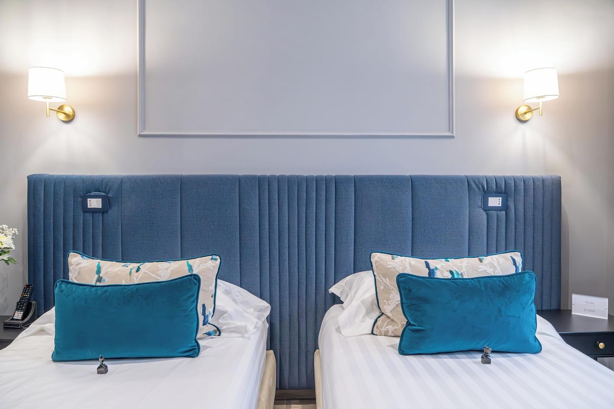 Una habitación de hotel con dos camas, cada una con un cojín azul turquesa y otro blanco con estampado floral. Hay lámparas fijadas a la pared a cada lado de las camas.