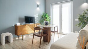 Uma sala de estar moderna com uma mesa redonda de vidro, duas cadeiras de madeira, um sofá, um armário de madeira, uma televisão e um vaso com planta.