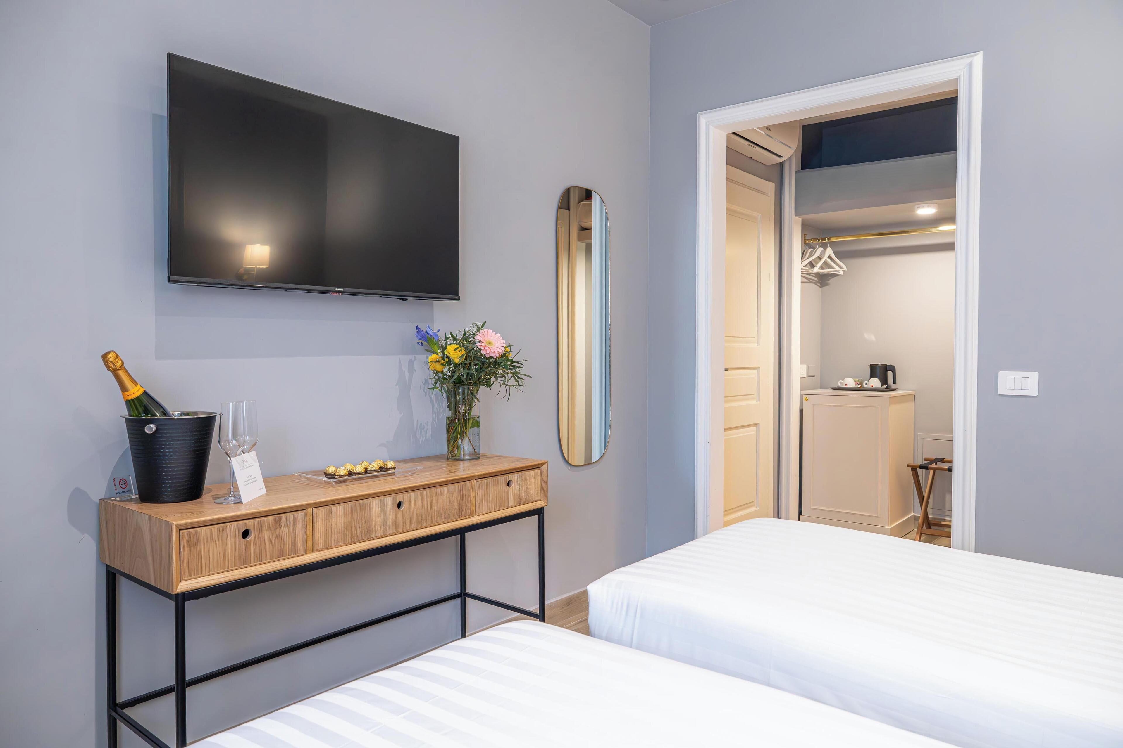 A hotel room with a TV, a wooden console table with a champagne bottle, glasses, and a vase of flowers. There are two beds and a mirror on the wall.