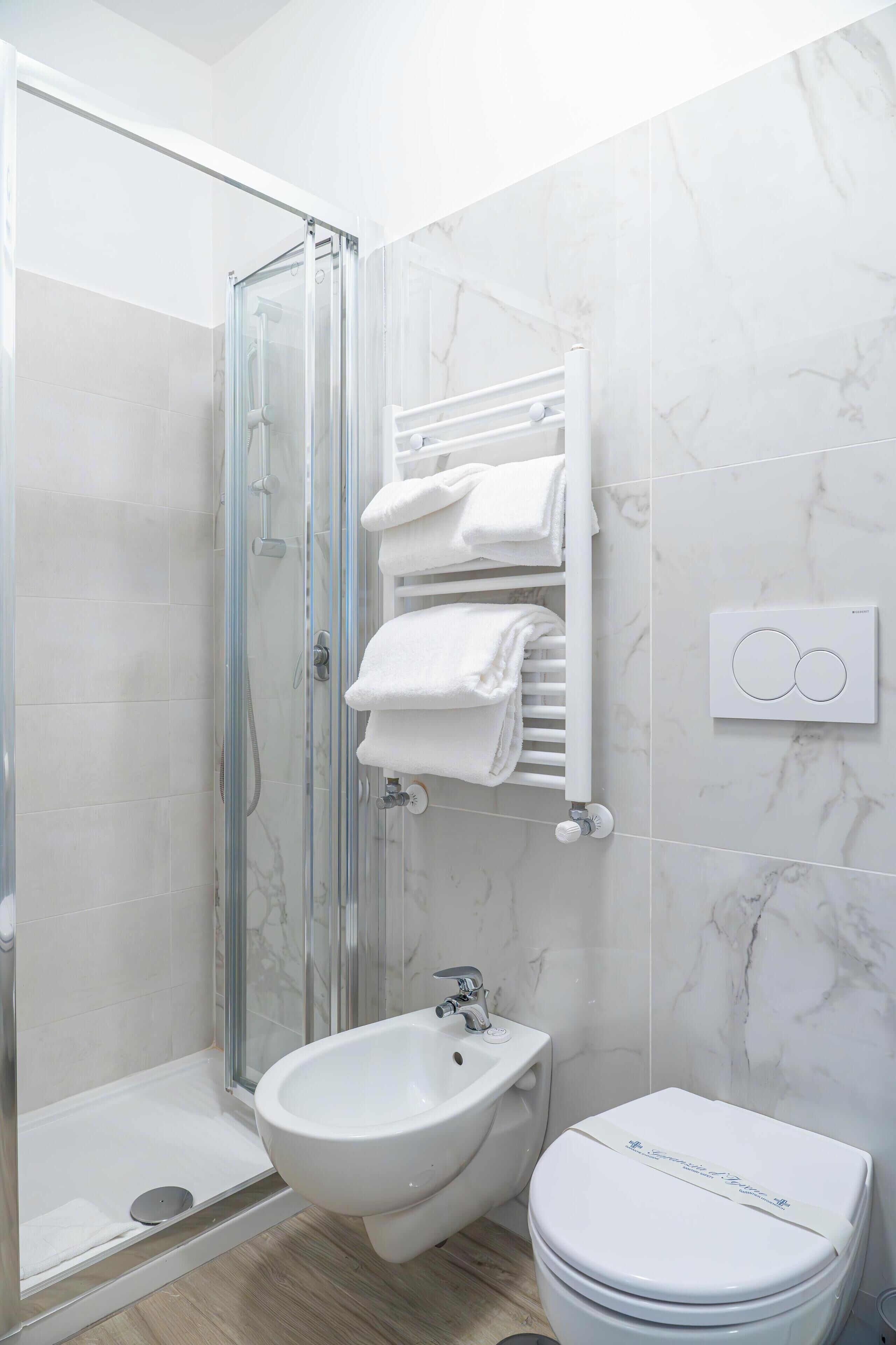 A modern bathroom with a glass shower enclosure, a bidet, a toilet, and a towel rack with neatly folded towels.