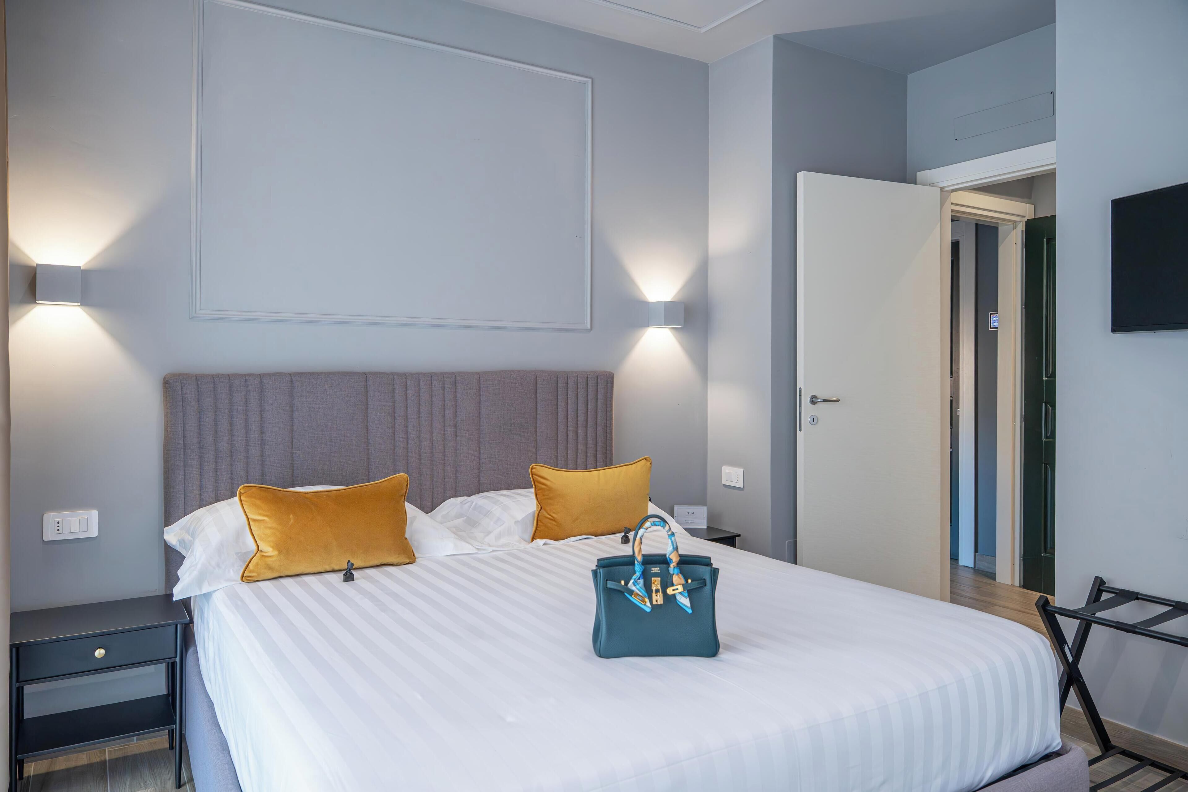 A neatly made bed with white bedding and two mustard pillows, a teal handbag on the bed, and a nightstand with a lamp.