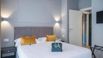 A neatly made bed with white bedding and two mustard pillows, a teal handbag on the bed, and a nightstand with a lamp.