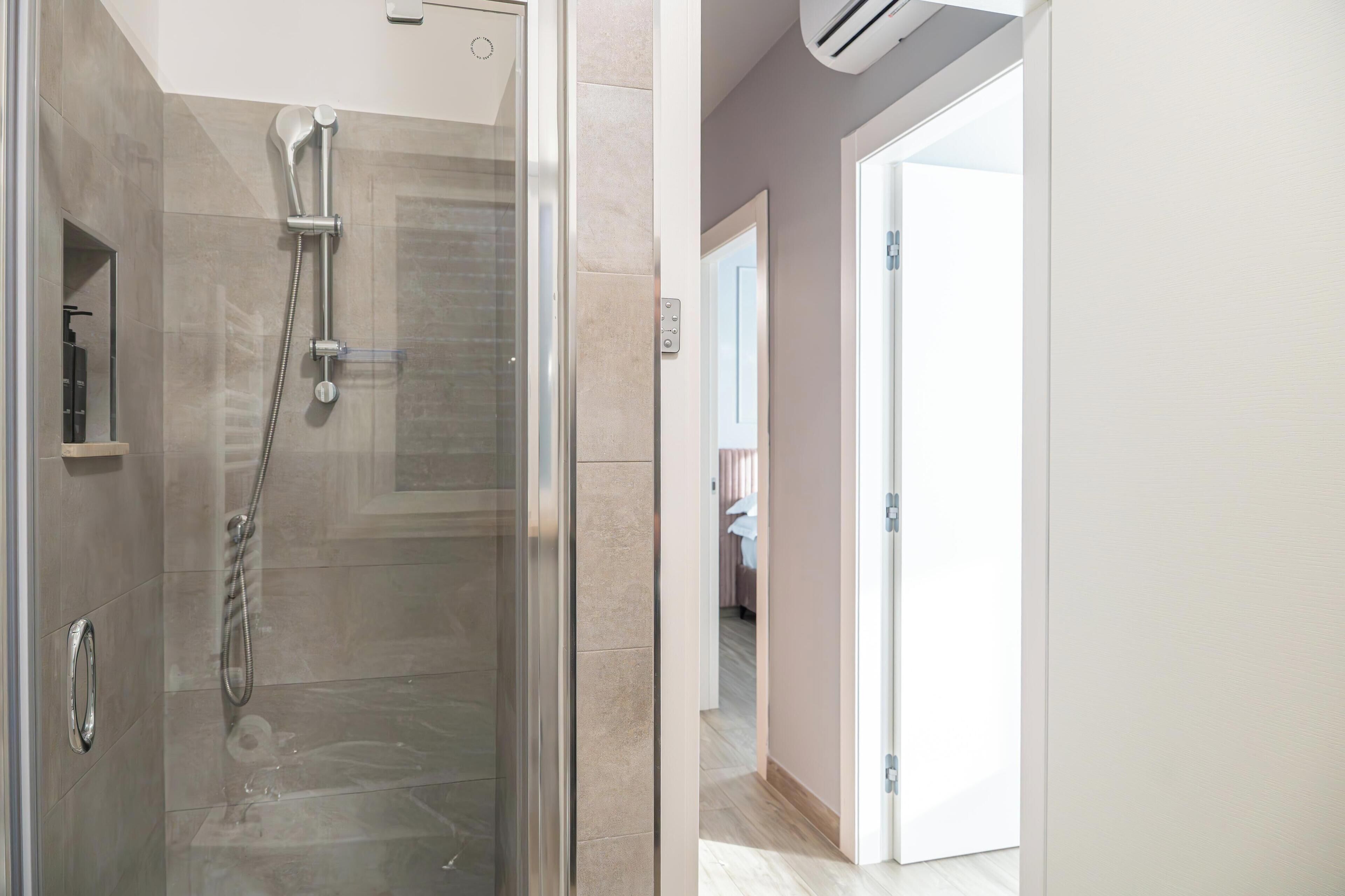 A modern bathroom with a glass-enclosed shower, a visible control panel, and a door leading to a bedroom.
