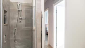 A modern bathroom with a glass-enclosed shower, a visible control panel, and a door leading to a bedroom.