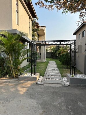 A gated entrance with a pathway leading to a building, surrounded by greenery and palm trees.