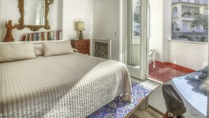 Un dormitorio con una cama grande, una mesita de noche de madera con una lámpara, una estantería y un espejo decorativo.