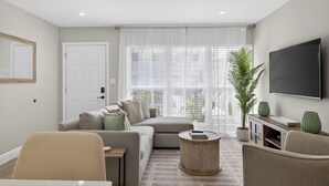 A modern living room with a sofa, armchairs, a coffee table, a TV, and a large window with blinds. - Coral Ridge Suites #1 - Heated Pool, Beach Gear (Fort Lauderdale)