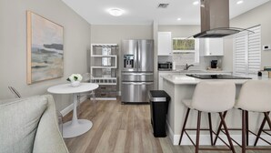 A modern kitchen with a stainless steel refrigerator, microwave, and stove, a white dining area with two bar stools, and a small round table with a vase of flowers. - Coral Ridge Suites #1 - Heated Pool, Beach Gear (Fort Lauderdale)