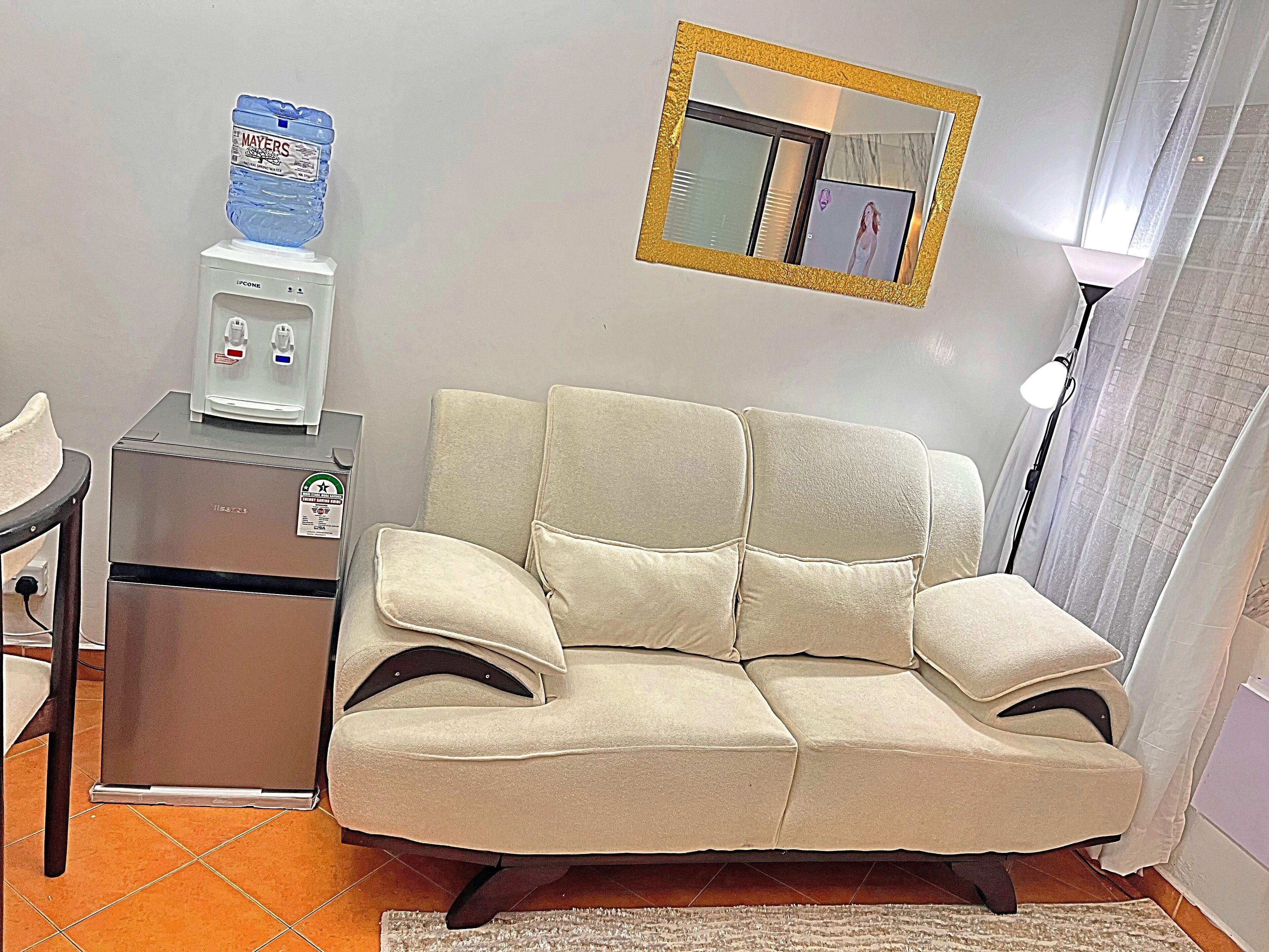 Un salon avec un canapé beige, un petit réfrigérateur, un distributeur d’eau et un miroir au mur.