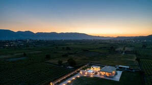 Vue aérienne d’un domaine viticole au crépuscule, avec une grande maison entourée de chemins et de champs bien éclairés.