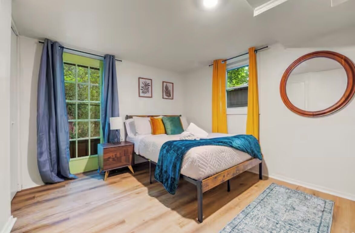 A bedroom with a bed, a nightstand, a window with curtains, a mirror, and a rug.