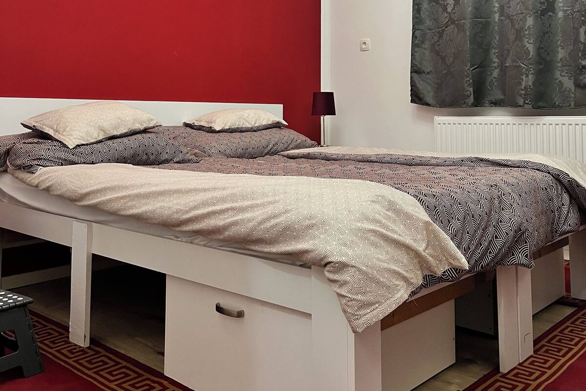 A single-bed room with a red wall, a patterned bedspread, a small stool, and a radiator.