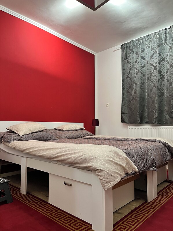 A single-bed room with a red wall, a patterned bedspread, a small stool, and a radiator.