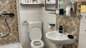 Ein Badezimmer mit Toilette, Waschbecken und Dusche. Es gibt Regale mit Pflegeprodukten und einen Spiegel über dem Waschbecken.