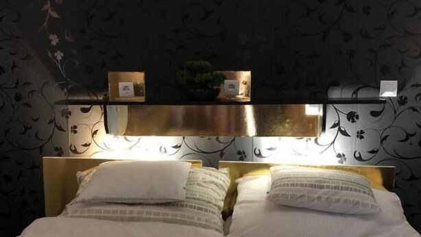 Ein ordentlich gemachtes Bett mit weißen Leinen und einer Streifen-Decke, ein Nachttisch mit Lampe sowie eine Wand mit floralem Muster.