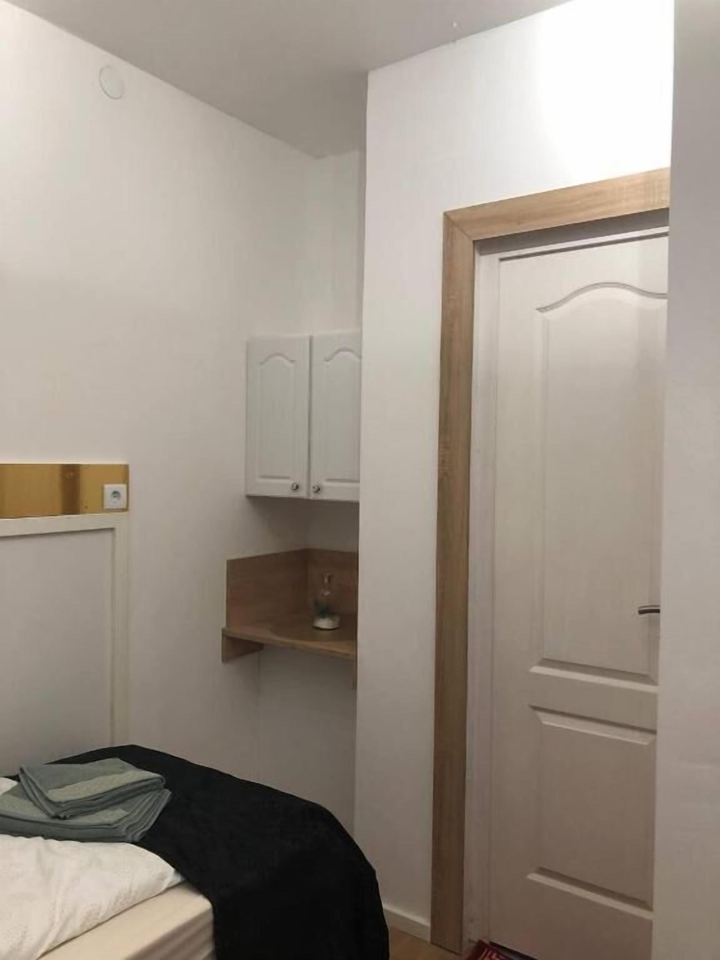 A small, sparsely furnished room with a bed, a wooden shelf, and a white door.