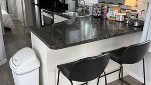 A modern kitchen with a granite countertop, stainless steel appliances, and a small island with bar stools. - Mira Bella South - 1BR Tiny Home in Lake Helen (Lake Helen)
