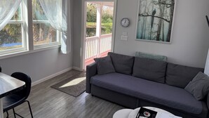 A living room with a grey sofa, a dining table with a black chair, a clock on the wall, and a window with sheer curtains. - Mira Bella South - 1BR Tiny Home in Lake Helen (Lake Helen)