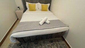 A neatly made bed with a grey mattress, white sheets, and two yellow pillows. A rolled white towel is placed on the bed. - Perfect Escapade (Ongata Rongai)