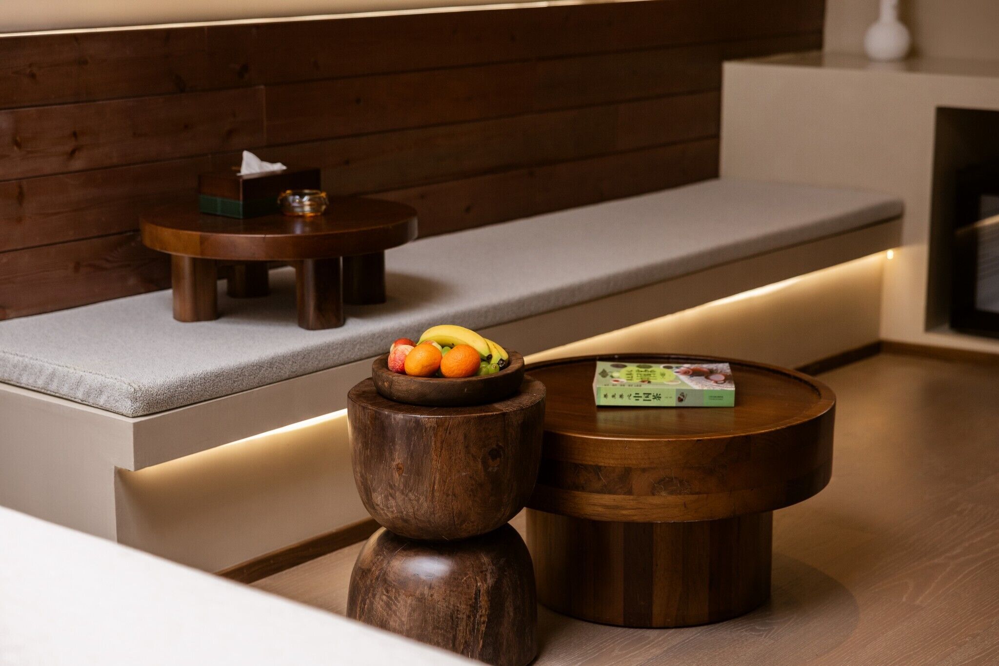 A modern seating area with a cushioned bench and two wooden tables, one with a book and the other with fruit.