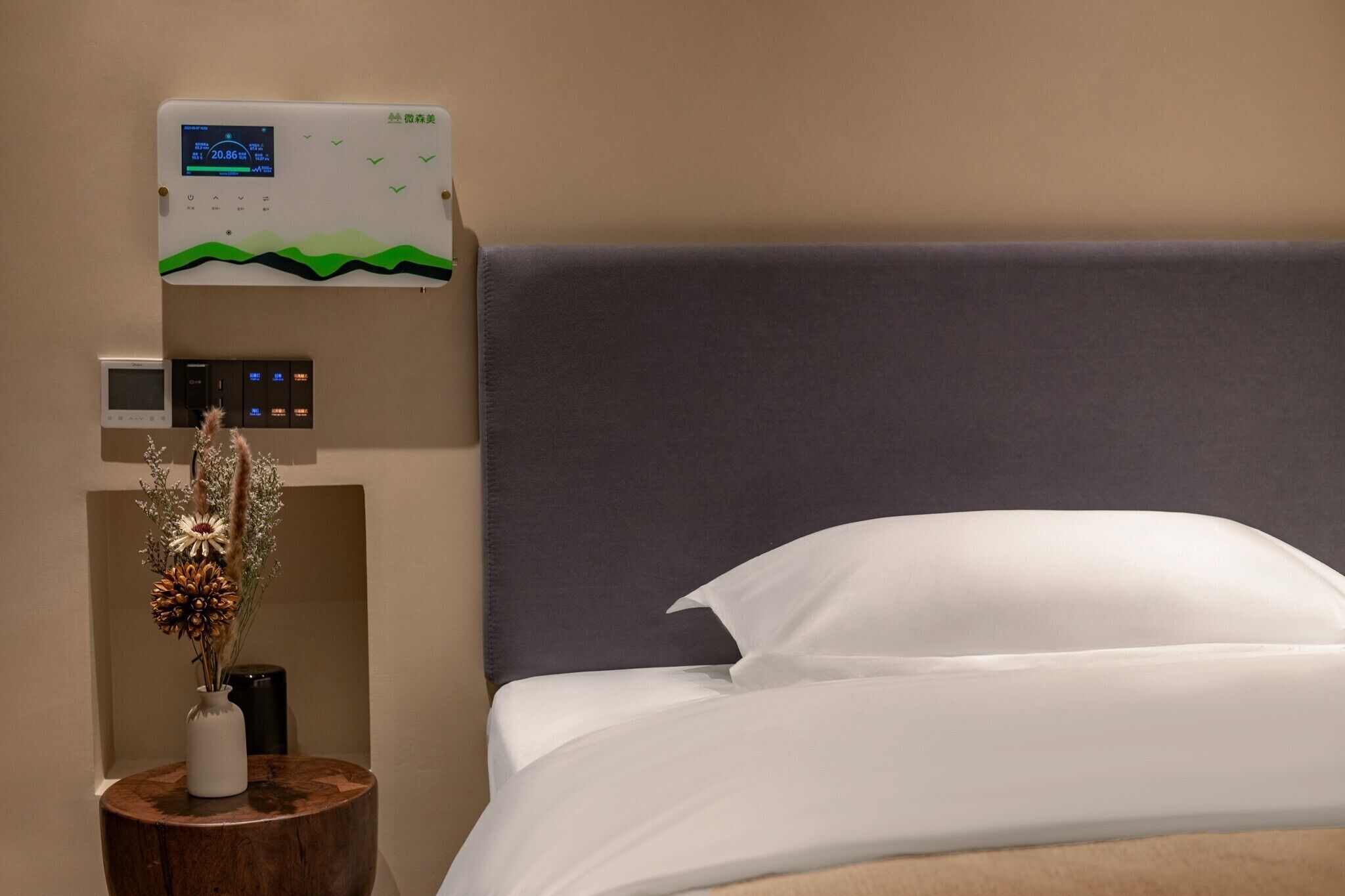 A hotel room with a bed, a bedside table with a vase of dried flowers, a thermostat, and a control panel.