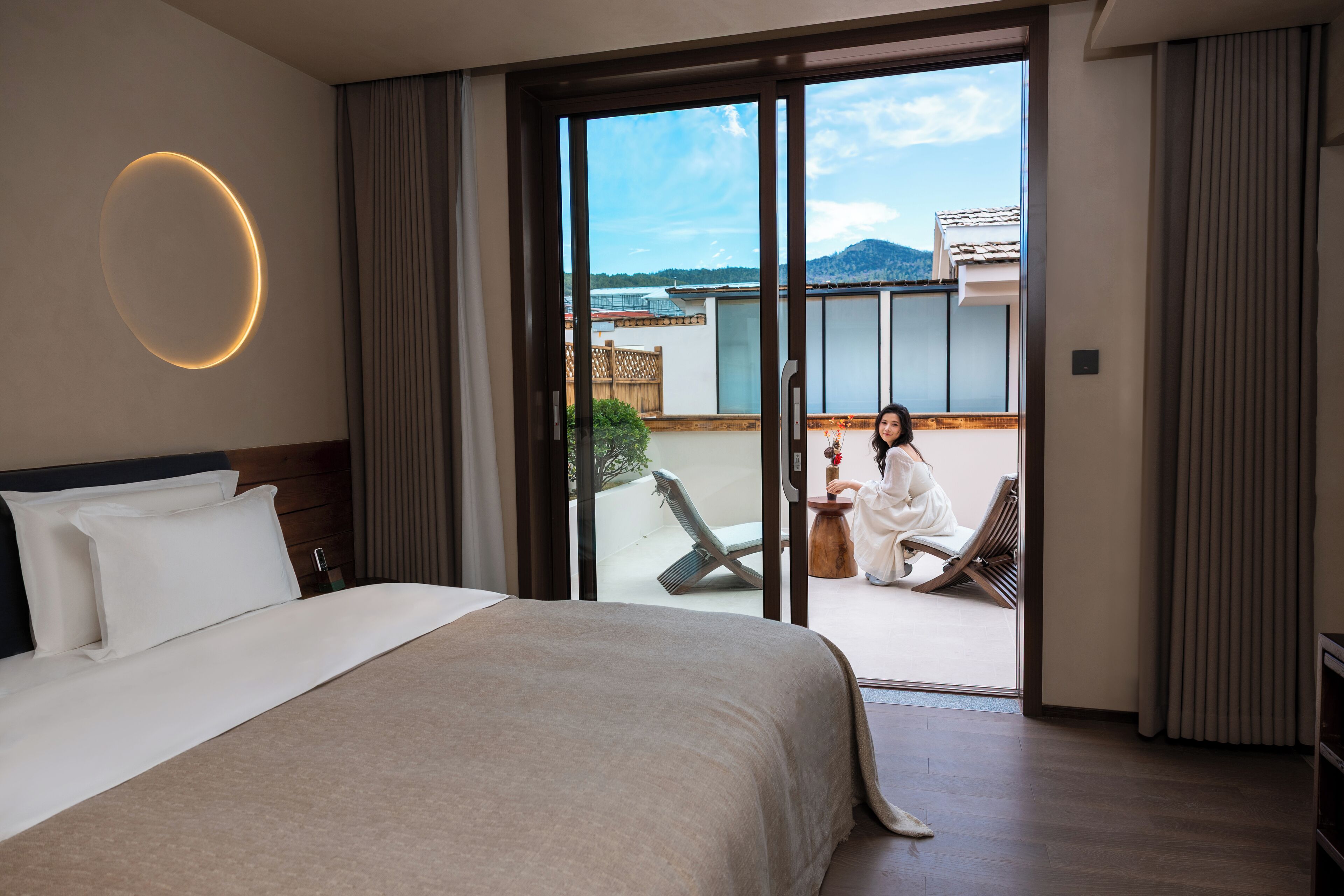 A hotel room with a large bed, a circular light fixture, a sliding glass door leading to an outdoor seating area with a view of the mountains.