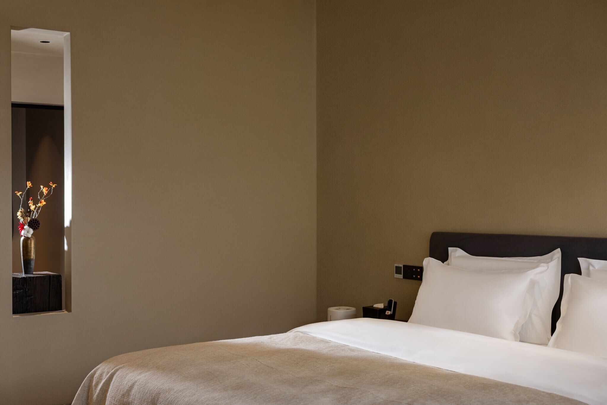 A hotel room with a bed, a bedside table, a mirror, and a wall-mounted light fixture.