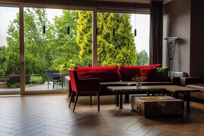 A modern living room with a red sofa, a wooden coffee table, and a view of lush greenery through large windows.