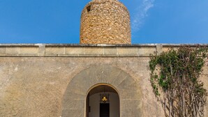 A stone building with a large arched entrance, a cylindrical tower, and a small garden area with plants and pots. - Villa Moli dén Llull (Sant Llorenç Des Cardassar)