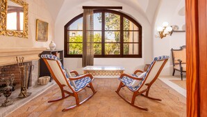 A room with a fireplace, two rocking chairs, a table, and a large arched window with curtains. - Villa Moli dén Llull (Sant Llorenç Des Cardassar)