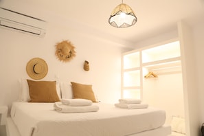 A hotel room with a bed, two pillows, a wall-mounted air conditioner, a decorative sun, and a window with a wooden hanger. - Villa Adelfia with private hot tub (Mykonos)
