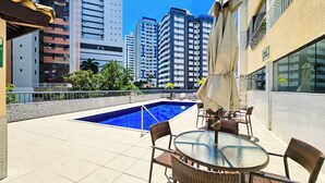 A rooftop pool area with a glass table and chairs, surrounded by high-rise buildings. - Apto c Piscina Academia e Churrasqueira em Pituba (Salvador)