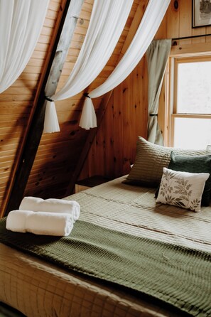 A cozy room with wooden walls, a bed with a green blanket, white pillows, and a window with curtains. - Northern MI Secluded A-Frame Cabin: ORVs, Fish/Hunt, Projector, Golf, EV, Pool (Grayling)