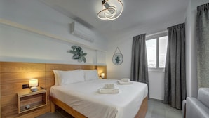 A hotel room with a wooden headboard, a large bed with white linens, a bedside table with a lamp, a wall-mounted air conditioner, and a window with curtains. - Central 1BR Studio Near St. Julian’s Hotspots w/AC (St. Julian`s)