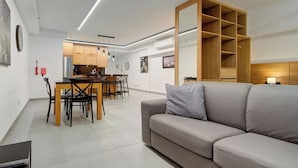 A modern living room with a grey sofa, wooden dining table, and chairs. There is a kitchen area with wooden cabinets and a built-in bookshelf. - Central 1BR Studio Near St. Julian’s Hotspots w/AC (St. Julian`s)