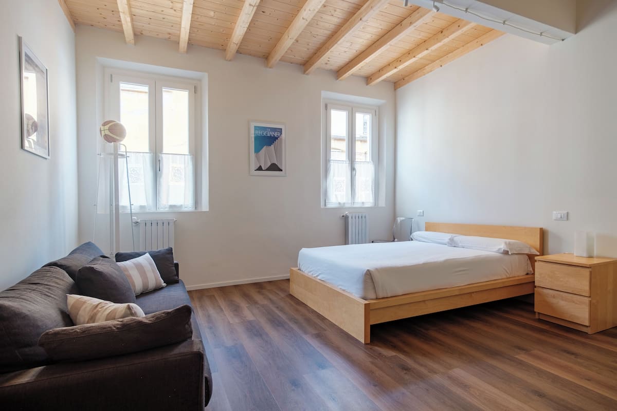 Une chambre à coucher avec un lit en bois, une table de chevet en bois, un canapé de couleur sombre et une fenêtre donnant sur un paysage agréable.