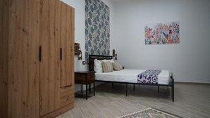A bedroom with a bed, a wooden wardrobe, a nightstand, a patterned rug, and a floral wall hanging. - Apartment   Ground Floor (balestrate)