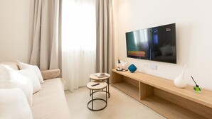 Habitación de hotel moderna con un televisor de pantalla plana grande montado en la pared, un sofá, una pequeña mesa redonda con un control remoto, una consola de madera con un objeto decorativo y una ventana con cortinas de gasa.