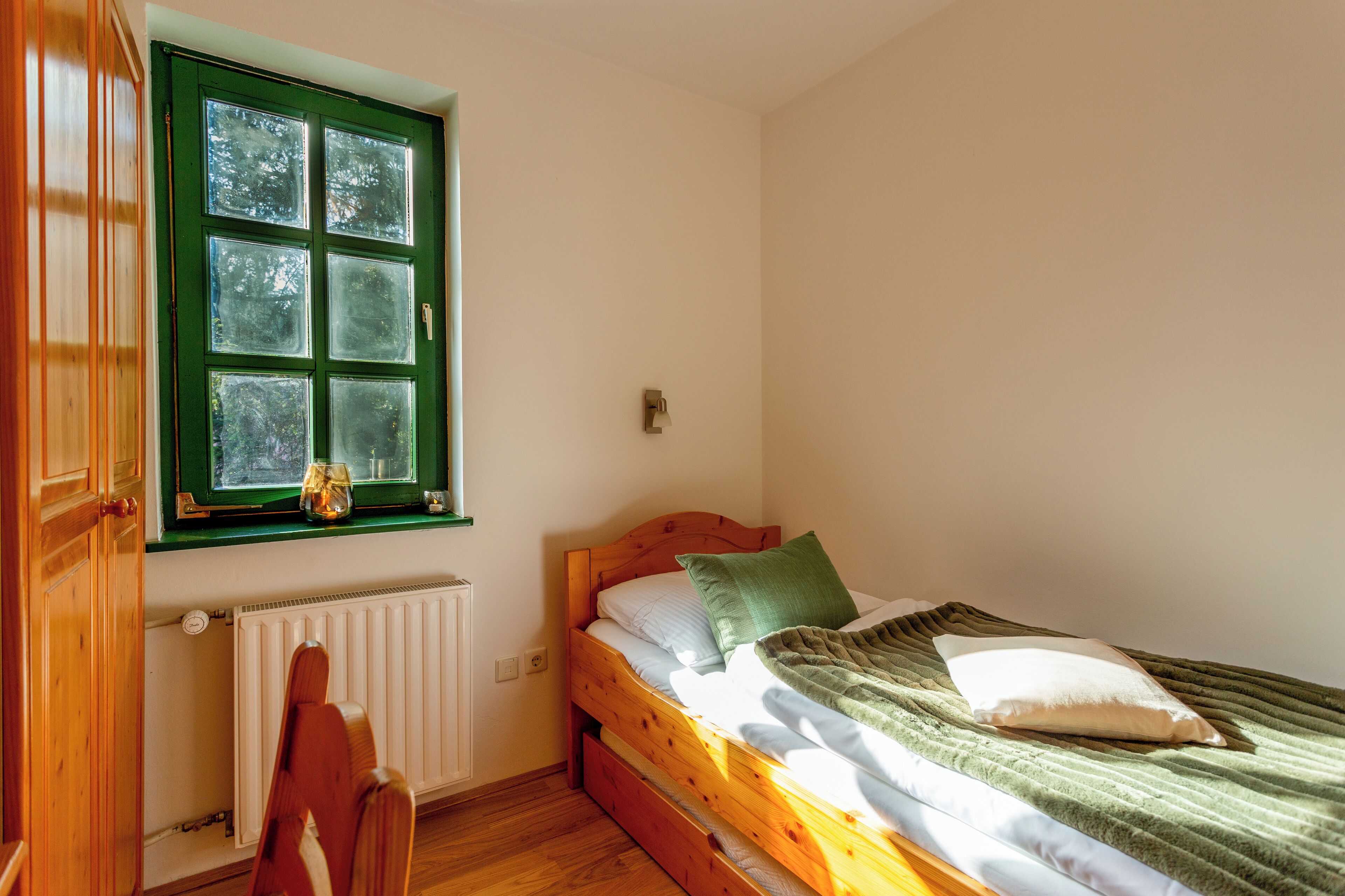 A small, single-bed room with a wooden headboard, a window with green shutters, and a radiator below.