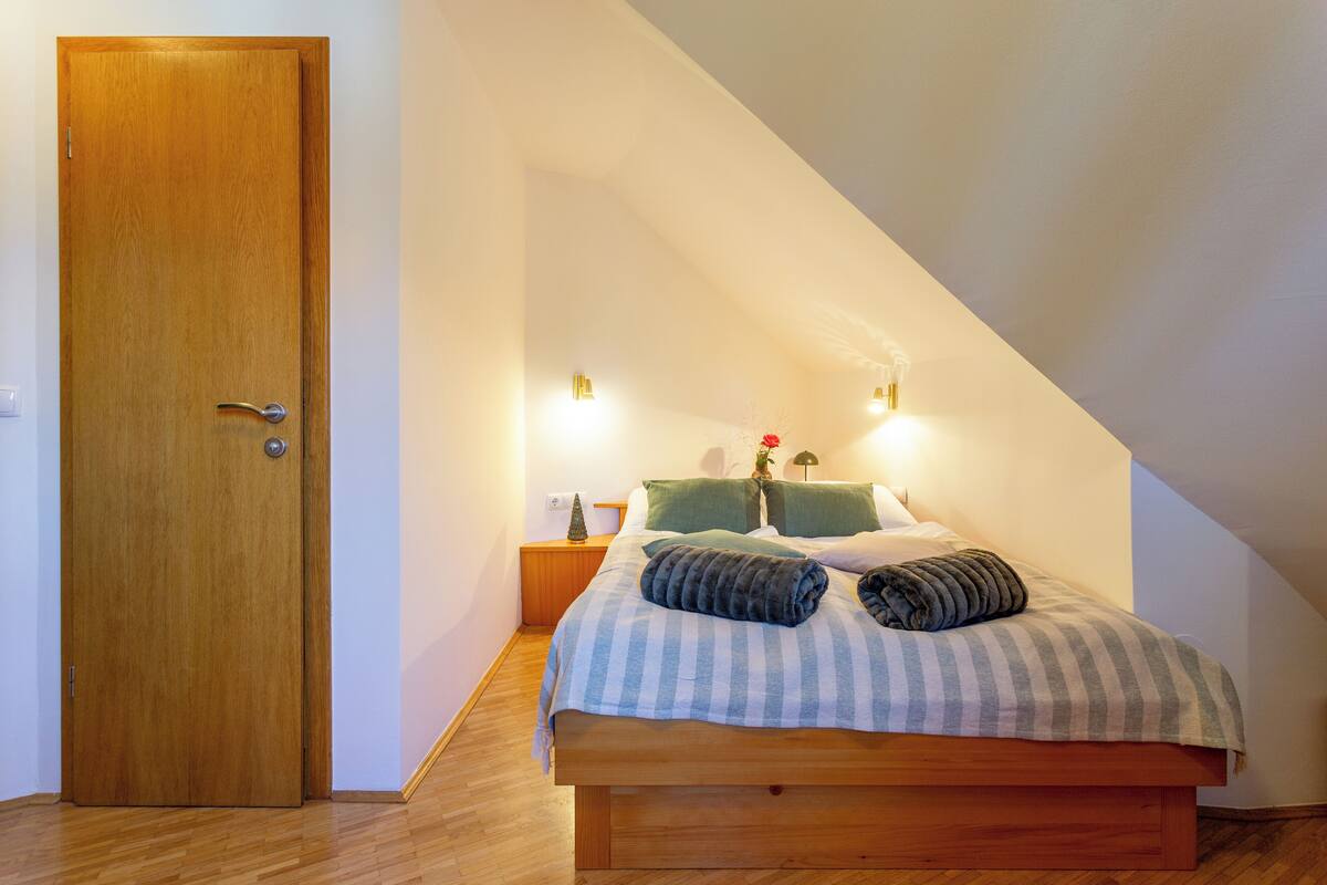A bedroom with a wooden door, a bed with a striped bedspread, two pillows, a nightstand with a lamp, and a small Christmas tree.