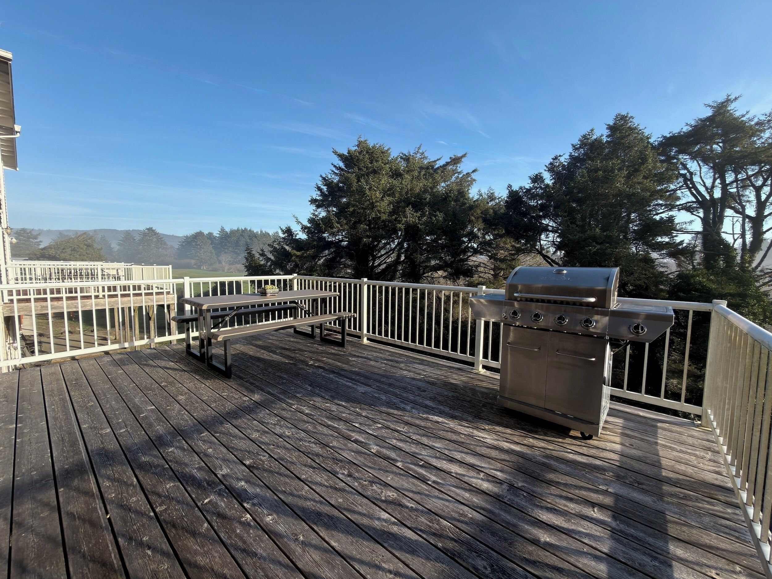 A wooden deck with a grill and picnic table, surrounded by a white railing and trees.