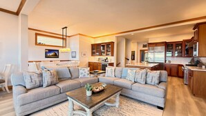 A spacious living room with a large sectional sofa, a wooden coffee table, and a dining area with white chairs. The kitchen features wooden cabinets and a large island. - Oceanfront with 4 Suites (Seaside)