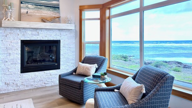 A living room with a fireplace, two patterned armchairs, a glass coffee table, and a large window offering a view of the ocean. - Oceanfront with 4 Suites (Seaside)