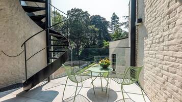 Un área de estar exterior moderna con una escalera en espiral, balaustrada de vidrio y vistas a árboles.