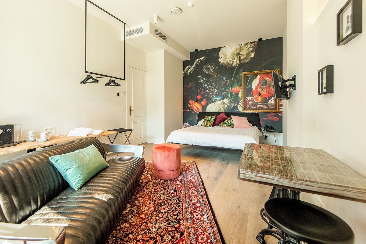 A modern hotel room with a sofa, a small table, a bed with colorful bedding, a large floral artwork on the wall, and a mirror.