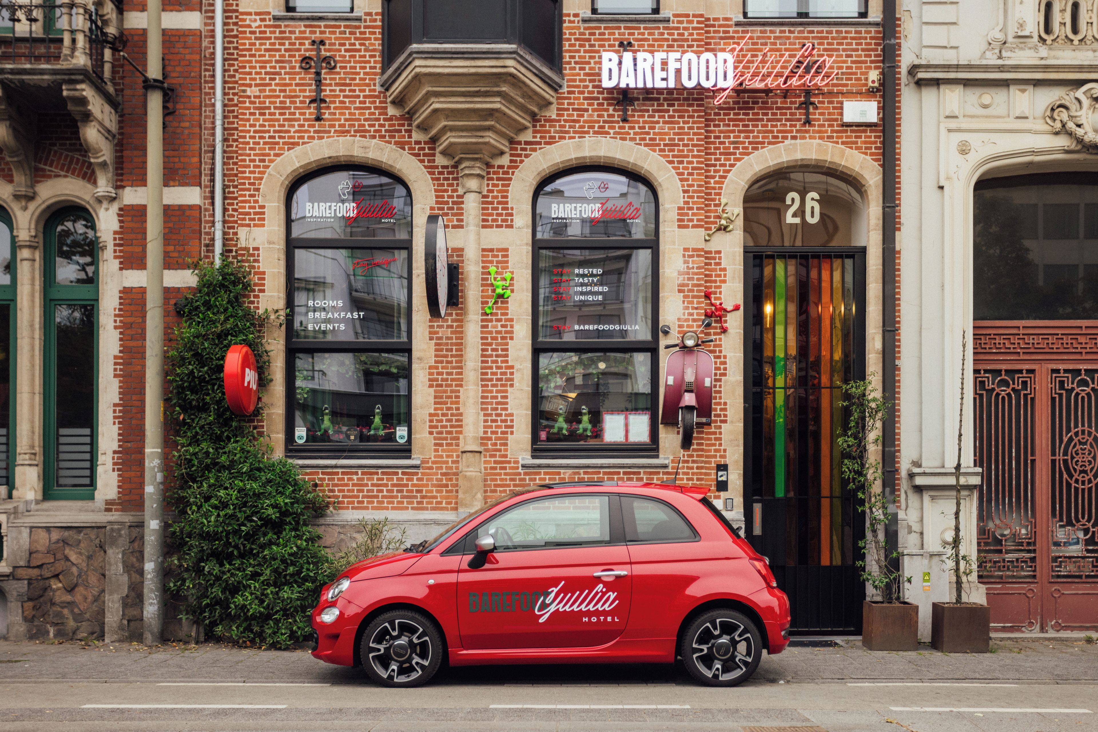 Een rode auto geparkeerd voor een hotel met een bakstenen gevel, grote gebogen ramen en een neonbord.