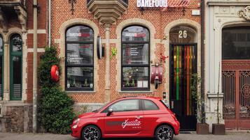 Een rode auto geparkeerd voor een hotel met een bakstenen gevel, grote gebogen ramen en een neonbord.