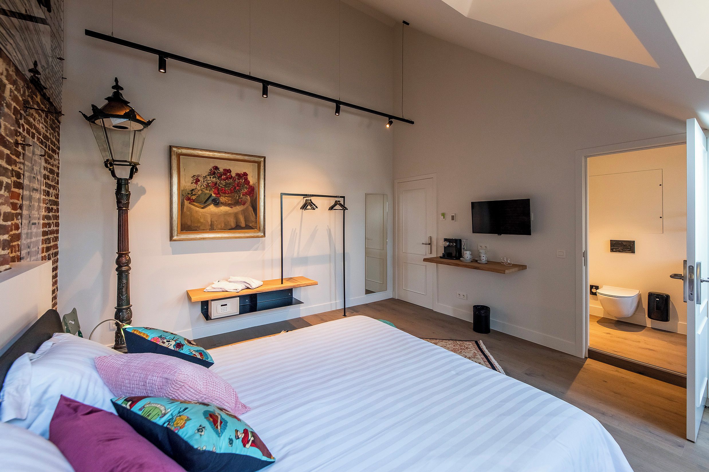 Ein modernes Schlafzimmer mit einem großen Bett, einem Nachttisch, einer Hängeleuchte, einem Gemälde, einem Kleiderschrank, einem Fernseher und einem durch eine offene Tür einsehbaren Badezimmer.