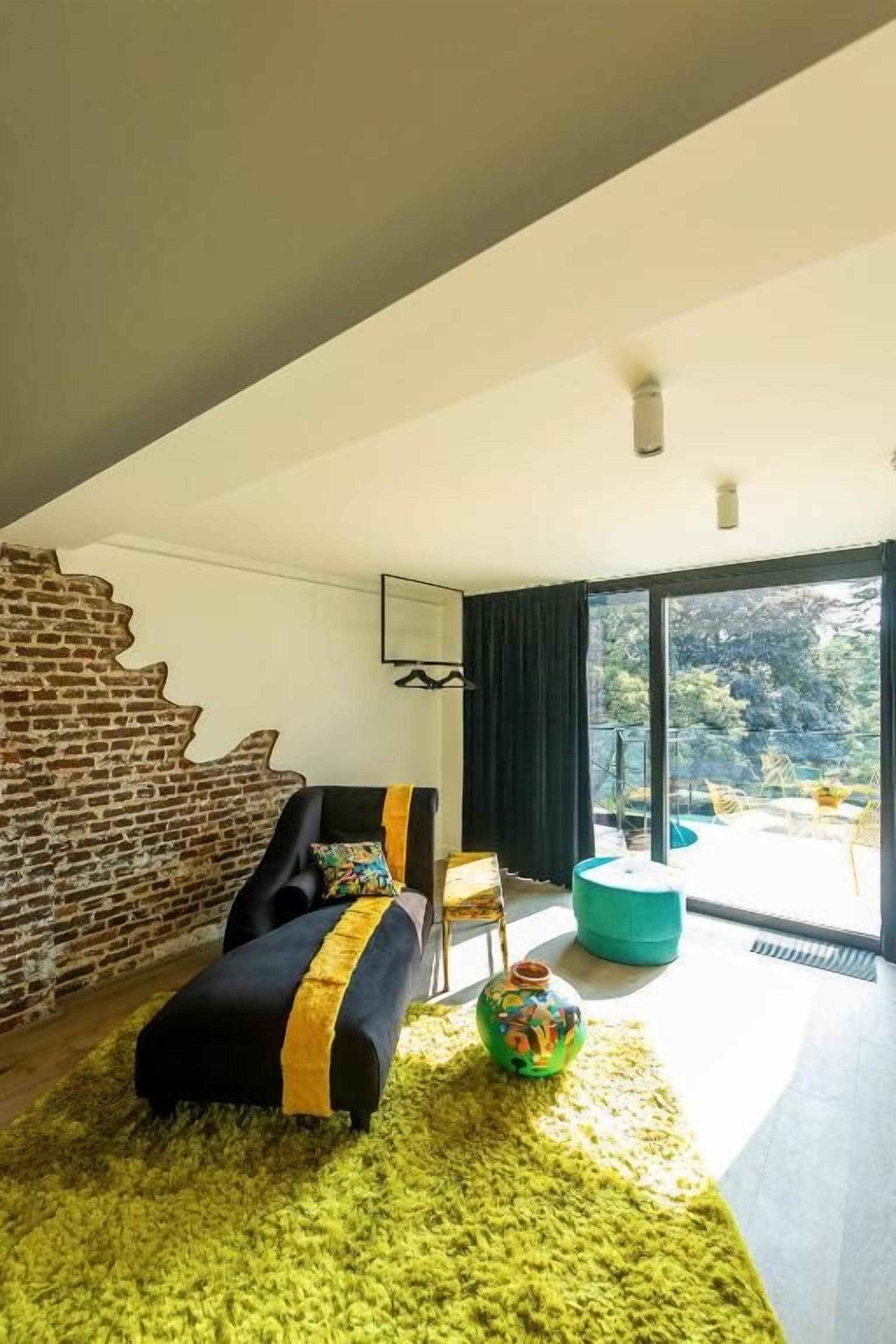 A modern living room with a brick wall, a black sofa with yellow accents, a green rug, and a large window offering a view of trees.