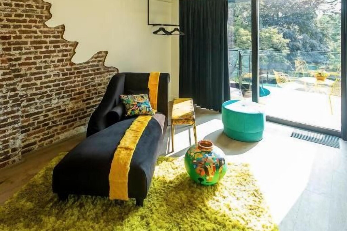 A modern living room with a brick wall, a black sofa with yellow accents, a green rug, and a large window offering a view of trees.